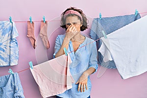 Young brunette woman doing laundry around string hangs smelling something stinky and disgusting, intolerable smell, holding breath