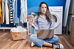 Young brunette woman doing laundry using laptop holding money in shock face, looking skeptical and sarcastic, surprised with open