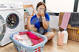 Young brunette woman doing laundry using smartphone smelling something stinky and disgusting, intolerable smell, holding breath