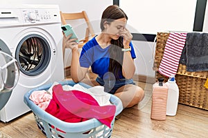 Young brunette woman doing laundry using smartphone tired rubbing nose and eyes feeling fatigue and headache