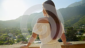 A young brunette woman, dressed in a flowing dress, stands on the villa terrace or balcony, enjoying mountain sunset