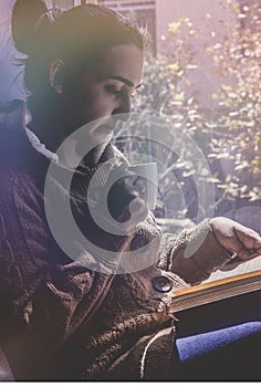 Young brunette woman drinking a cup of tea or coffee and reading a book. Female sitting at home by the window and read a book