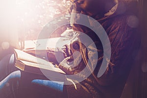 Young brunette woman drinking a cup of tea or coffee and reading a book. Female sitting at home by the window and read a book