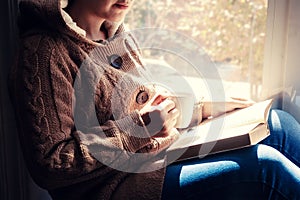 Young brunette woman drinking a cup of tea or coffee and reading a book. Female sitting at home by the window and read a book