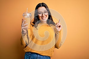 Young brunette woman drinking fresh orange juice from take away bottle over yellow background surprised with an idea or question