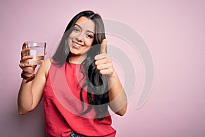 Young brunette woman drinking a glass of fresh water over pink isolated background happy with big smile doing ok sign, thumb up