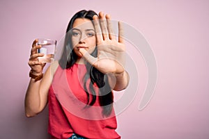 Young brunette woman drinking a glass of fresh water over pink isolated background with open hand doing stop sign with serious and