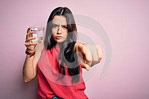 Young brunette woman drinking a glass of fresh water over pink isolated background pointing with finger to the camera and to you,
