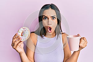 Young brunette woman eating doughnut and drinking coffee afraid and shocked with surprise and amazed expression, fear and excited