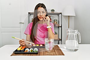 Young brunette woman eating sushi using chopsticks mouth and lips shut as zip with fingers