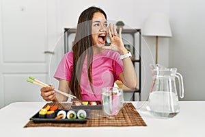 Young brunette woman eating sushi using chopsticks shouting and screaming loud to side with hand on mouth