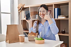 Young brunette woman eating take away food at home showing smartphone screen smiling happy and positive, thumb up doing excellent