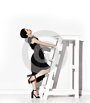 Young brunette woman in elegant black dress and shoes standing posing at big white ladder with her head thrown back