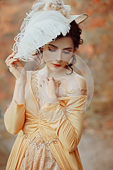 A young brunette woman with an elegant, hairstyle in a hat with a strass feathers. Lady in a yellow vintage dress walks