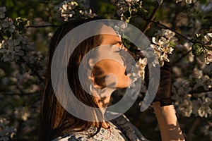 Young woman enjoys by blooming apple trees and sniffs it flowers with closed eyes in spring garden. Allergy concept.