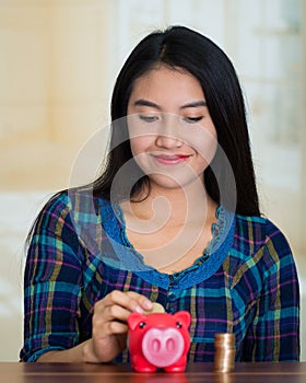 Young brunette woman facing camera, holding piggy bank and placing money inside it, smiling happily