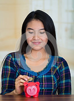 Young brunette woman facing camera, holding piggy bank and placing money inside it, smiling happily