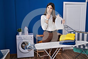 Young brunette woman with folded laundry after ironing covering mouth with hand, shocked and afraid for mistake