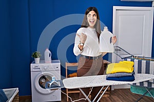 Young brunette woman with folded laundry after ironing pointing thumb up to the side smiling happy with open mouth