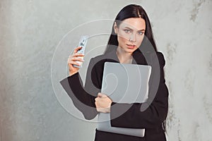 Young brunette woman in formal black suit holding laptop and wad of dollars