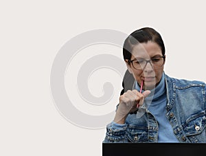 A young Caucasian brunette woman in a denim jacket and glasses with a pensive look holds a pen in her hands. Free space