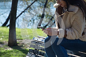 Young brunette woman with glasses, sitting on top of a bench, in a park, checking social networks on her cell phone. Concept