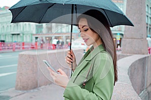 Young brunette woman in a green jacket on a walk. A beautiful girl under an umbrella uses a smartphone