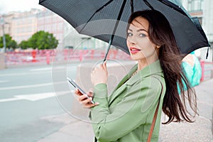 Young brunette woman in a green jacket on a walk. A beautiful girl under an umbrella uses a smartphone