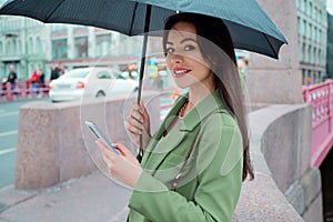 Young brunette woman in a green jacket on a walk. A beautiful girl under an umbrella uses a smartphone