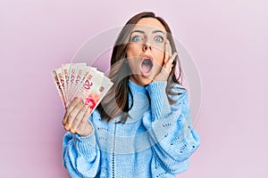 Young brunette woman holding 20 israel shekels banknotes afraid and shocked, surprise and amazed expression with hands on face