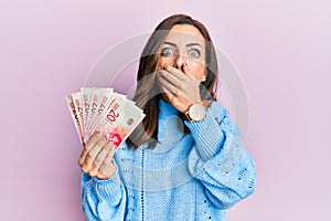Young brunette woman holding 20 israel shekels banknotes shocked covering mouth with hands for mistake