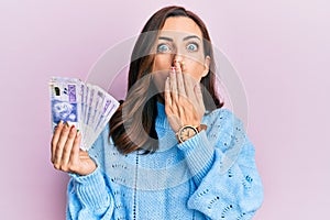 Young brunette woman holding 20 swedish krona banknotes covering mouth with hand, shocked and afraid for mistake