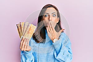 Young brunette woman holding 500 norwegian krone banknotes covering mouth with hand, shocked and afraid for mistake