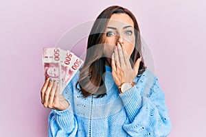 Young brunette woman holding 500 swedish krona banknotes covering mouth with hand, shocked and afraid for mistake