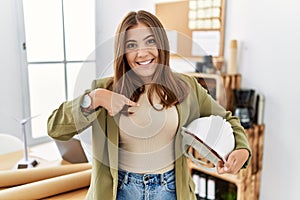 Young brunette woman holding architect hardhat at the office pointing finger to one self smiling happy and proud