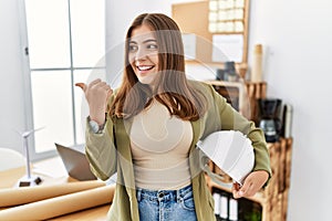 Young brunette woman holding architect hardhat at the office pointing thumb up to the side smiling happy with open mouth