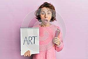 Young brunette woman holding art notebook and colored pencils afraid and shocked with surprise and amazed expression, fear and
