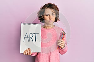 Young brunette woman holding art notebook and colored pencils relaxed with serious expression on face