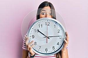 Young brunette woman holding big clock covering face afraid and shocked with surprise and amazed expression, fear and excited face