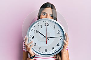 Young brunette woman holding big clock covering face in shock face, looking skeptical and sarcastic, surprised with open mouth
