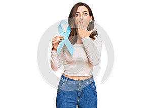 Young brunette woman holding blue ribbon covering mouth with hand, shocked and afraid for mistake