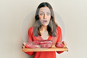 Young brunette woman holding board with raw meat afraid and shocked with surprise and amazed expression, fear and excited face