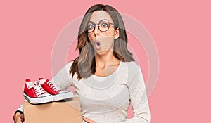 Young brunette woman holding box with red casual shoes scared and amazed with open mouth for surprise, disbelief face