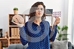 Young brunette woman holding brain and pills as mental health concept puffing cheeks with funny face