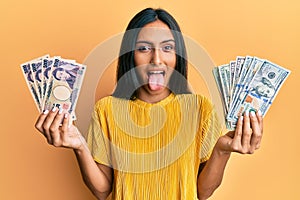 Young brunette woman holding bunch of dollars and yens sticking tongue out happy with funny expression