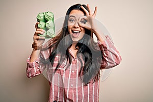 Young brunette woman holding cardboard cup of fresh raw eggs over isolated background with happy face smiling doing ok sign with