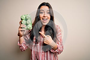 Young brunette woman holding cardboard cup of fresh raw eggs over isolated background very happy pointing with hand and finger