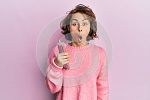 Young brunette woman holding colored pencils scared and amazed with open mouth for surprise, disbelief face