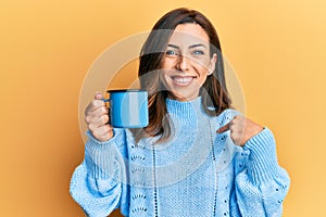 Young brunette woman holding a cup of coffee pointing finger to one self smiling happy and proud
