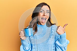 Young brunette woman holding a cup of coffee pointing thumb up to the side smiling happy with open mouth